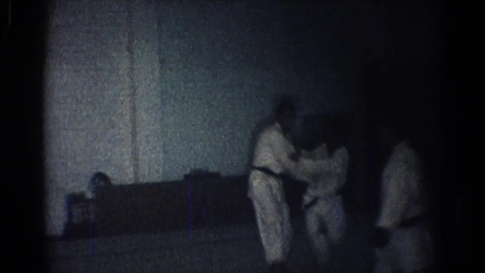 In low-light 1966 San Gabriel footage, men in white karate gi practice a close defensive hold while another person stands nearby.
