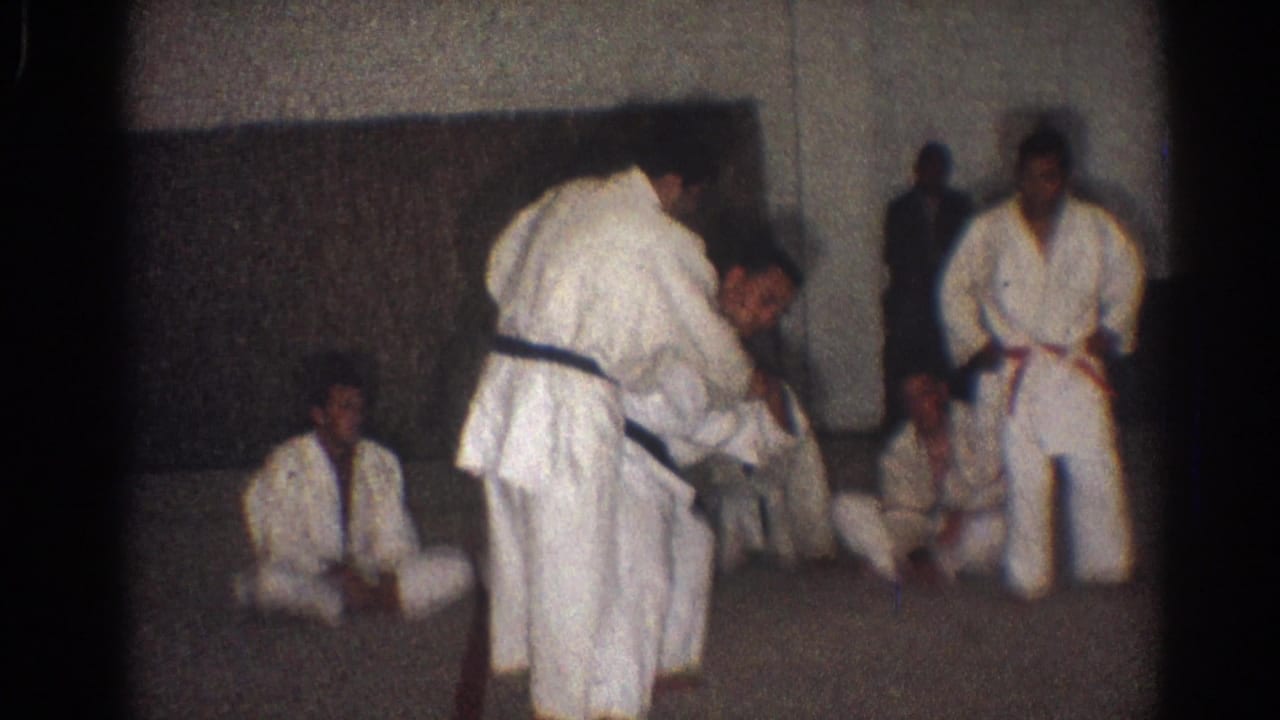 A man in a white gi bends over a partner during a karate demonstration as seated and standing observers watch in the San Gabriel training room.