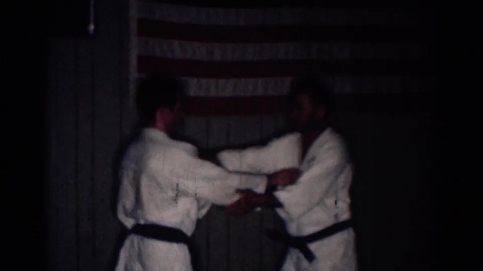 Two men in white gi practice facing holds in a dim dojo, with an American flag hanging on the wall behind them.
