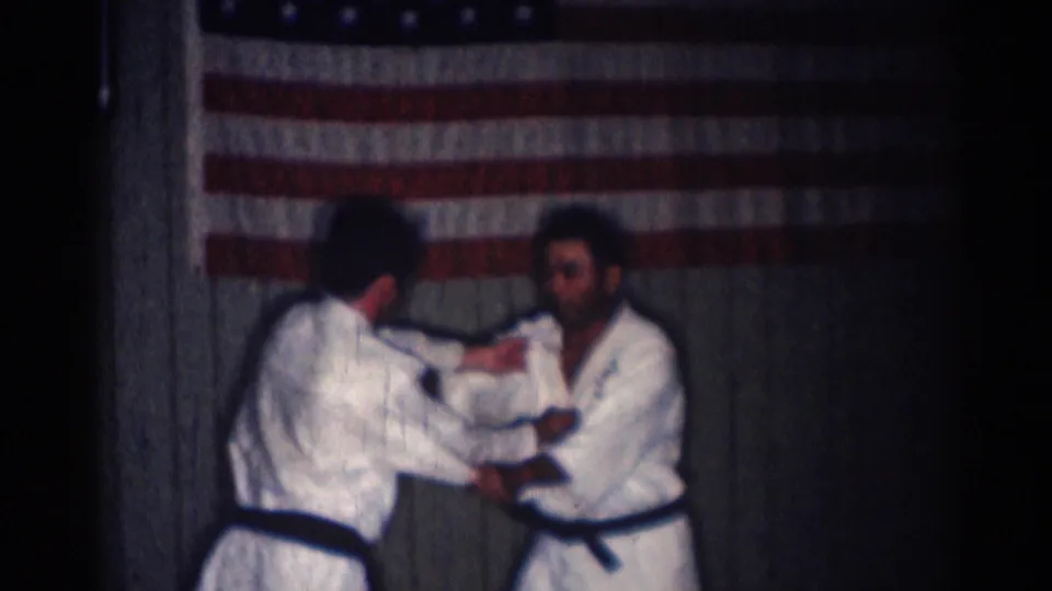 In 1966 San Gabriel footage, two men in white martial arts uniforms practice a close-contact technique before an American flag.