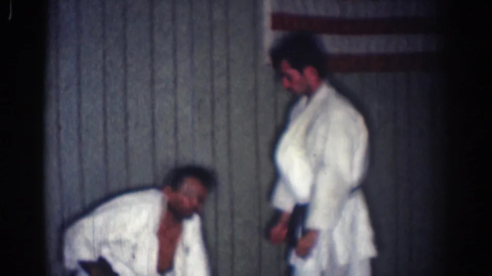 A standing man in a white gi pauses as his partner crouches low after the throw in the San Gabriel martial arts footage.