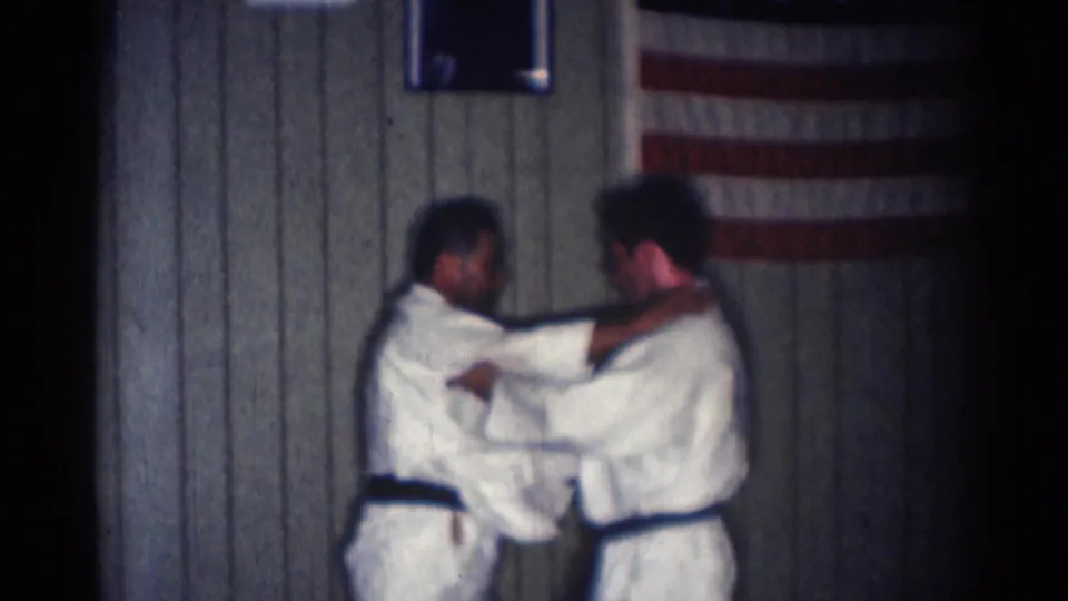 Two men in white gi hold each other at close range during a martial arts drill in the 1966 San Gabriel interior.