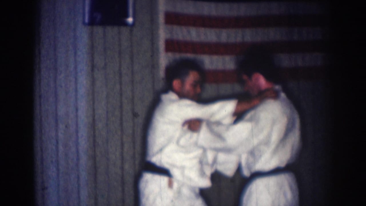 Two men in white karate uniforms grapple in front of a wood-paneled wall and American flag.