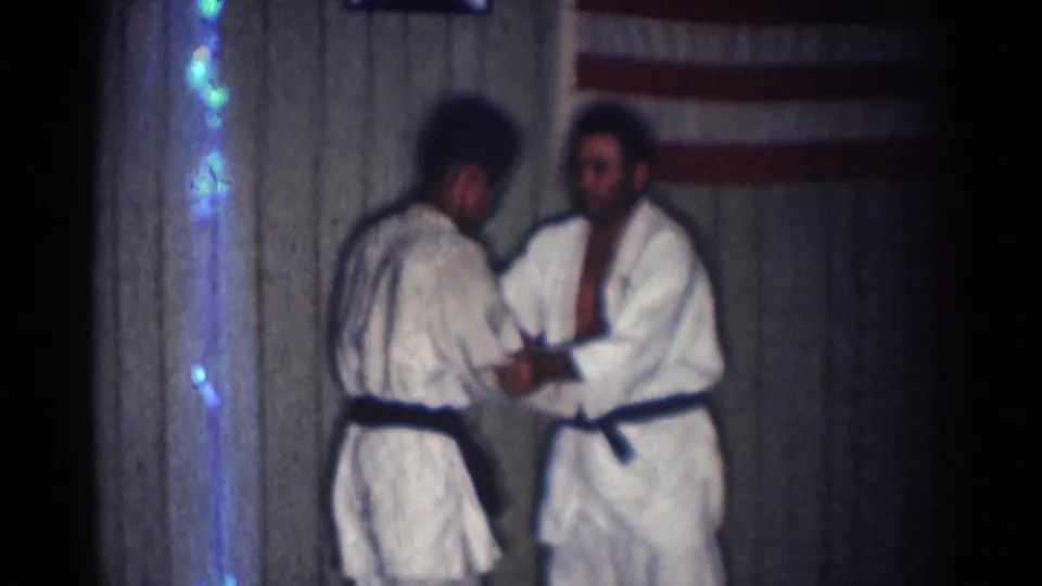 Two adult men in white karate uniforms and black belts reset or discuss positioning in the San Gabriel practice room near an American flag.