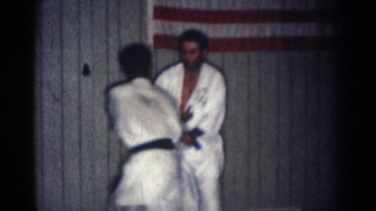 Two men in white gi face each other in a dojo with an American flag behind them.