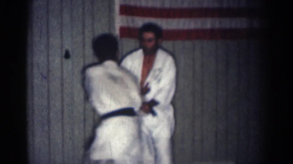 In 1966 San Gabriel, California, two adult black-belt martial artists in white gi face each other at the start of a practice exchange inside a dojo.