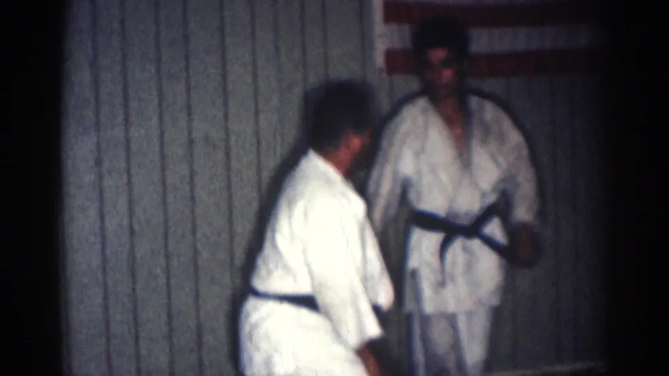 Two adult martial artists in white gi stand near the wall after the throw sequence, with one figure closer to the camera and the other facing forward.