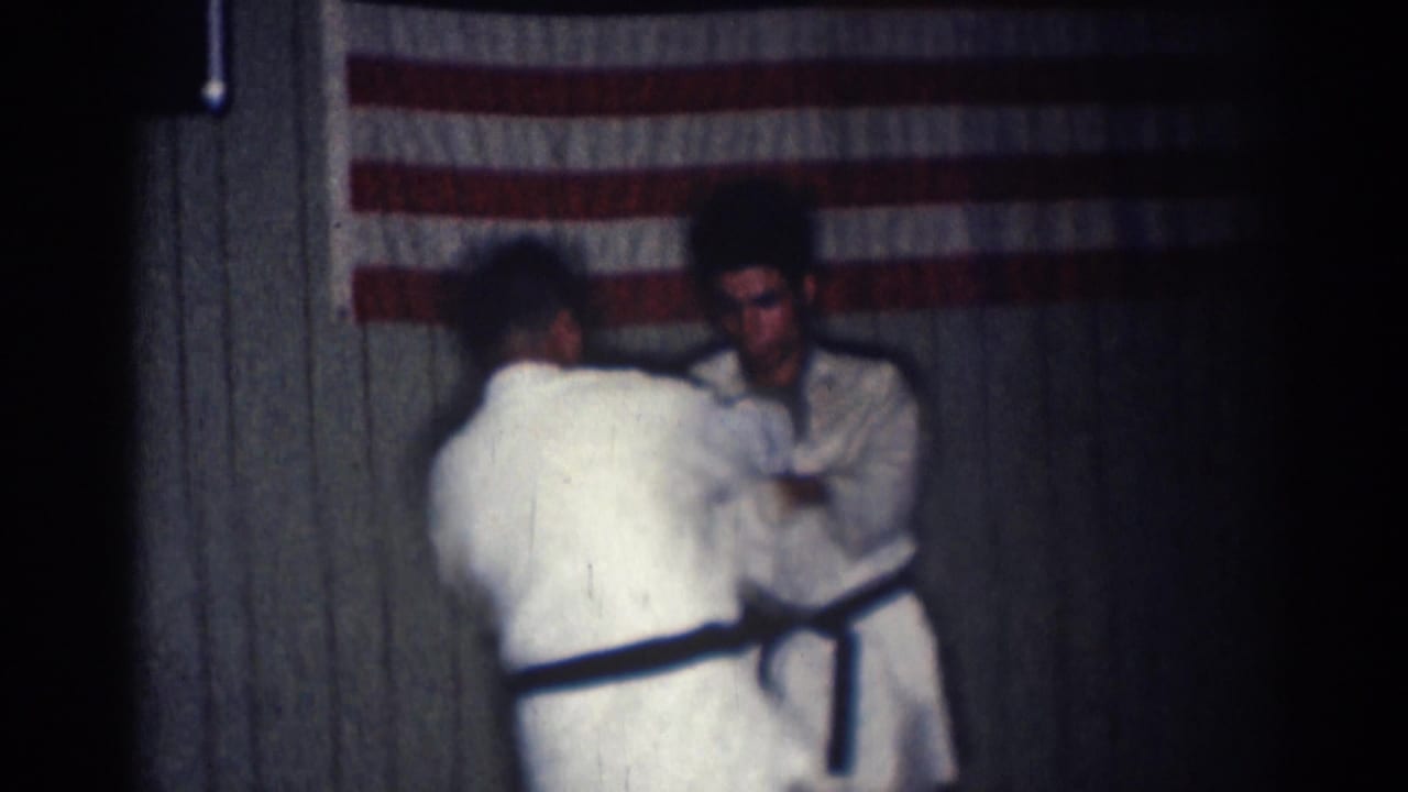 Two men in white gi and black belts practice near a wall with an American flag.