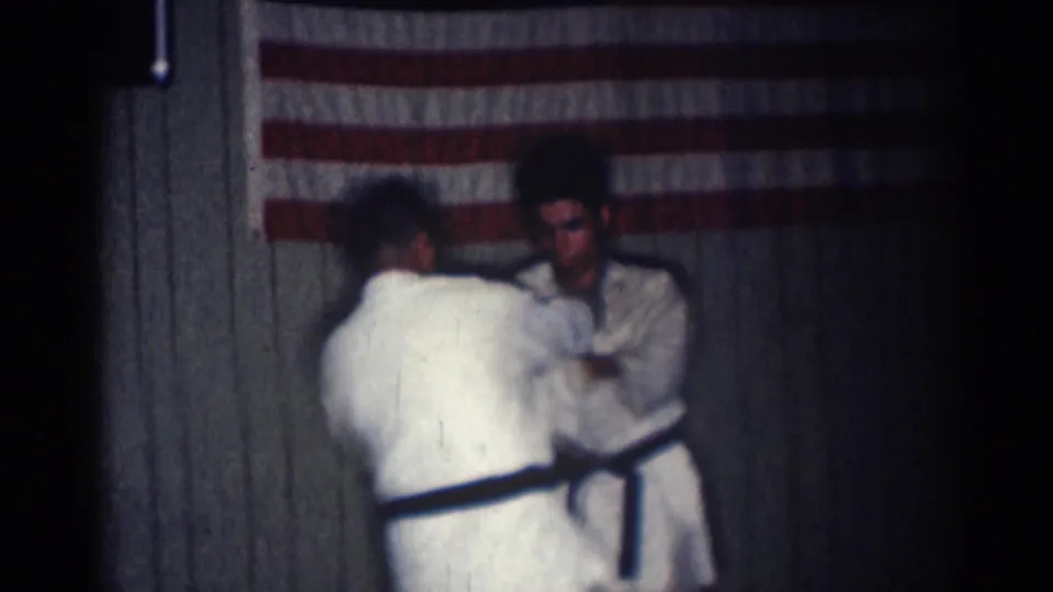 In San Gabriel, California, 1966, two adult men in white martial arts uniforms face each other on a dark mat beneath an American flag.