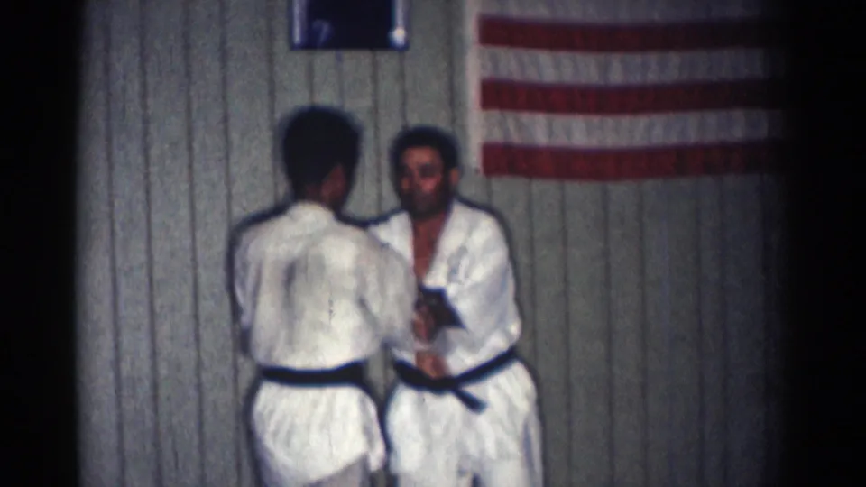 The two black-belt martial artists face each other again in the indoor San Gabriel practice space, with the American flag partly visible behind them.