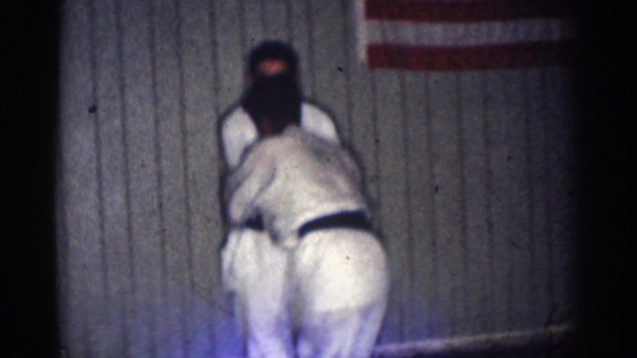 Two men in white karate uniforms grapple in front of a wood-paneled wall and American flag.