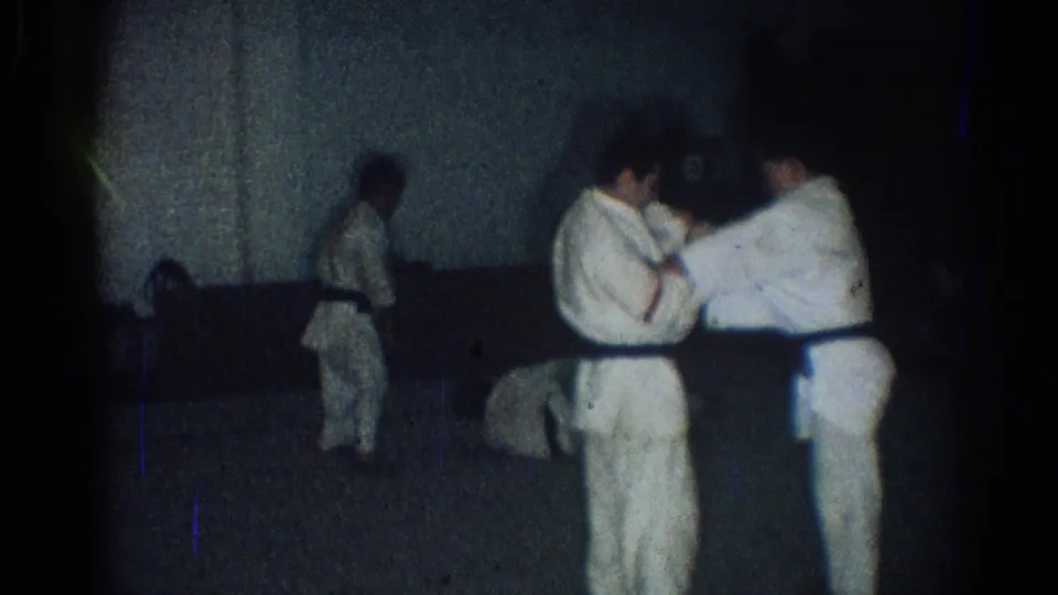 Multiple karate practitioners are visible in the gym, with two men in white gi working a close-contact technique in the foreground.