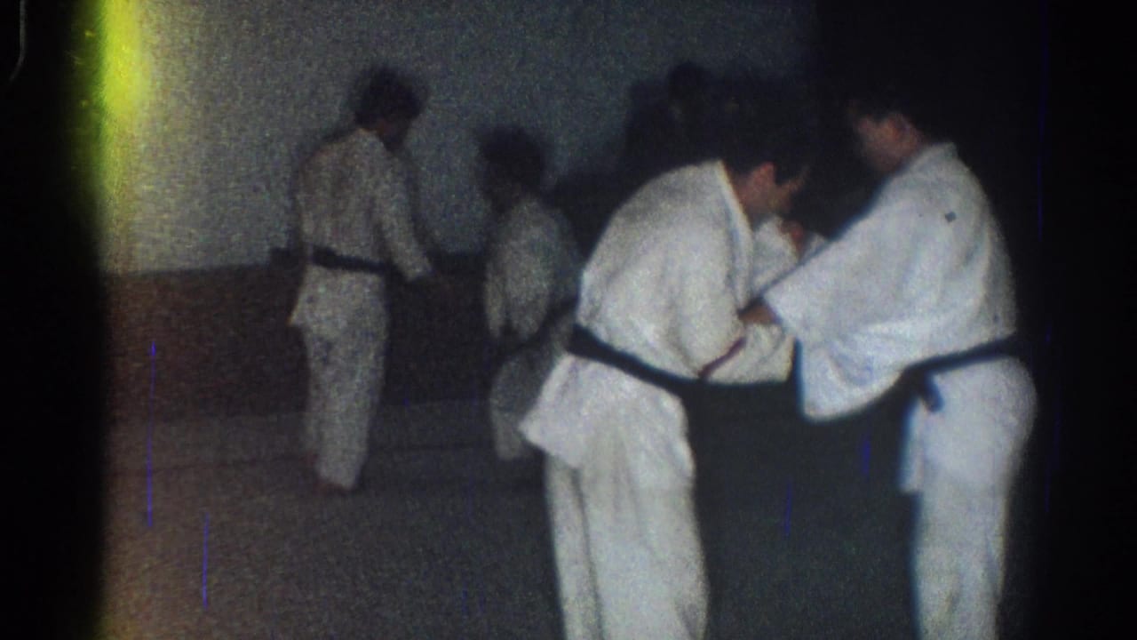 Several adults in white gi stand on a dark mat in a dim room while two grapple in front.