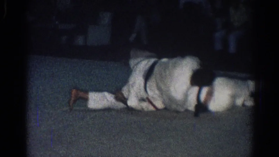 Wide low-light frame from the 1966 San Gabriel footage showing two martial arts practitioners extended on the mat in a ground position.