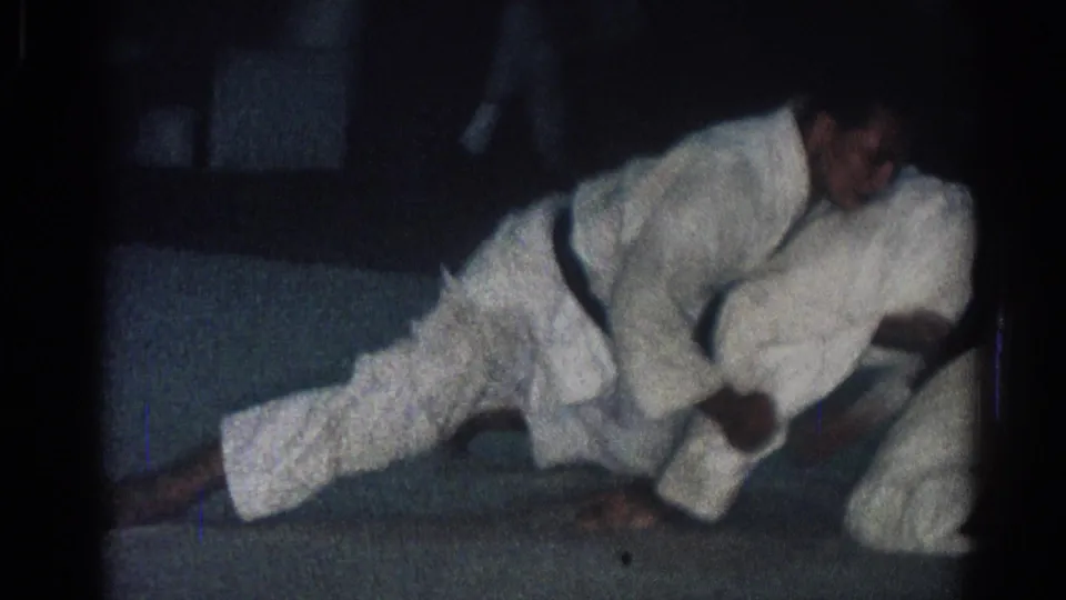 1966 San Gabriel 8mm frame showing two adult men in white judo gis grappling near the floor on a dim indoor mat.