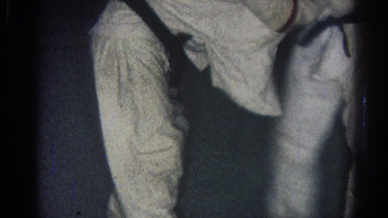 1966 San Gabriel home-movie frame showing two men in white gi and black belts grappling in a dark indoor room.
