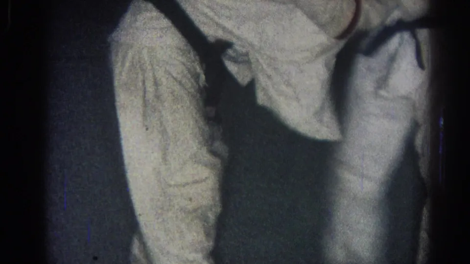 1966 San Gabriel home-movie frame showing two men in white gi and black belts grappling in a dark indoor room.
