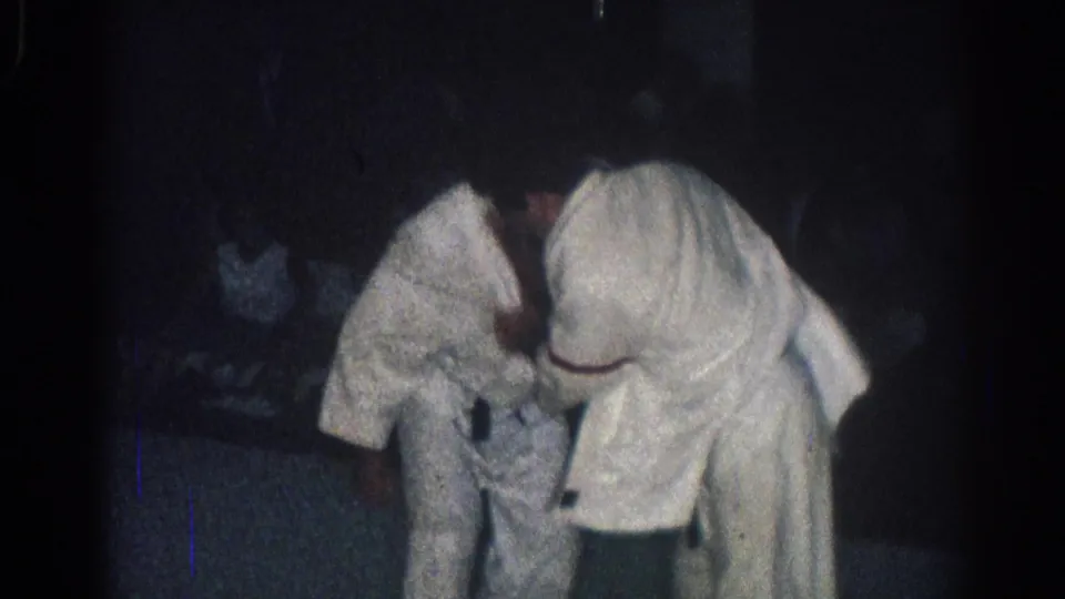 A 1966 San Gabriel home-movie frame shows two men in white gi and black belts grappling with spectators faintly behind them.