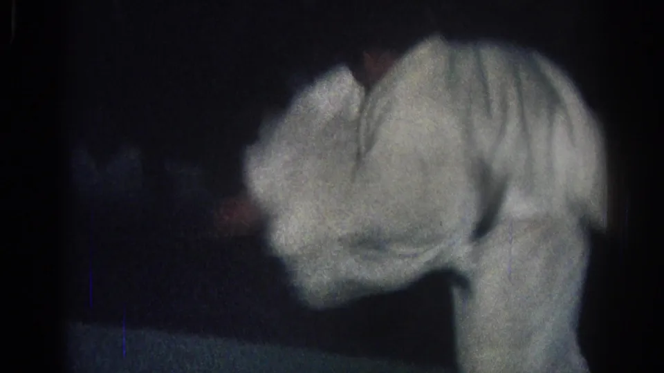 In 1966 San Gabriel footage, two martial artists in white gi are partially blurred as they bend and grapple in a dark indoor training space.