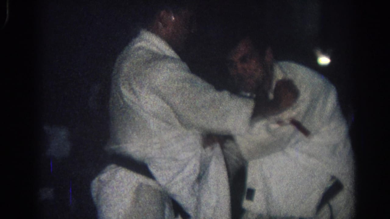 Two people in white gi grapple closely, one leaning across the other in a dark room.