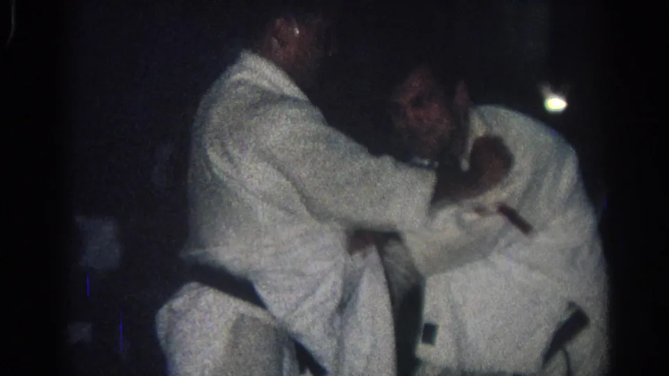 Two judo practitioners in white gi grapple at close range in the dim San Gabriel training room, with faces partly visible and motion blur.