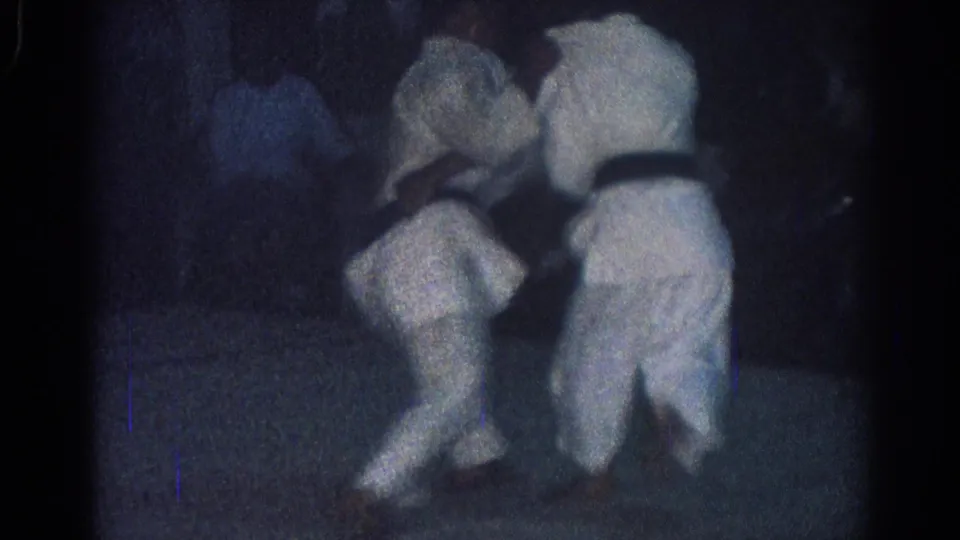 In a dark 1966 San Gabriel dojo scene, one practitioner crouches low while holding another, suggesting a judo throw or takedown in progress.
