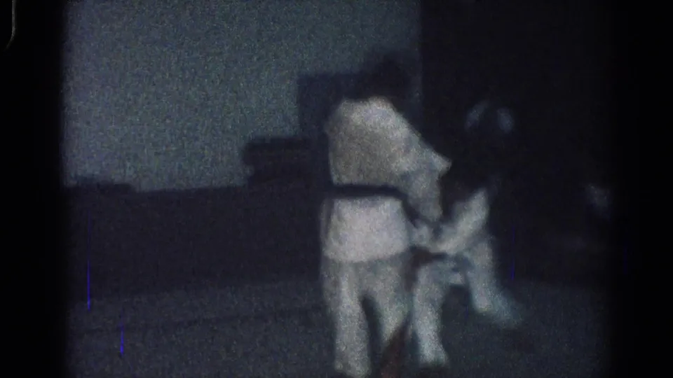 Two men in white judo gi appear to grapple during a 1966 San Gabriel indoor practice session, filmed in low light on 8mm.