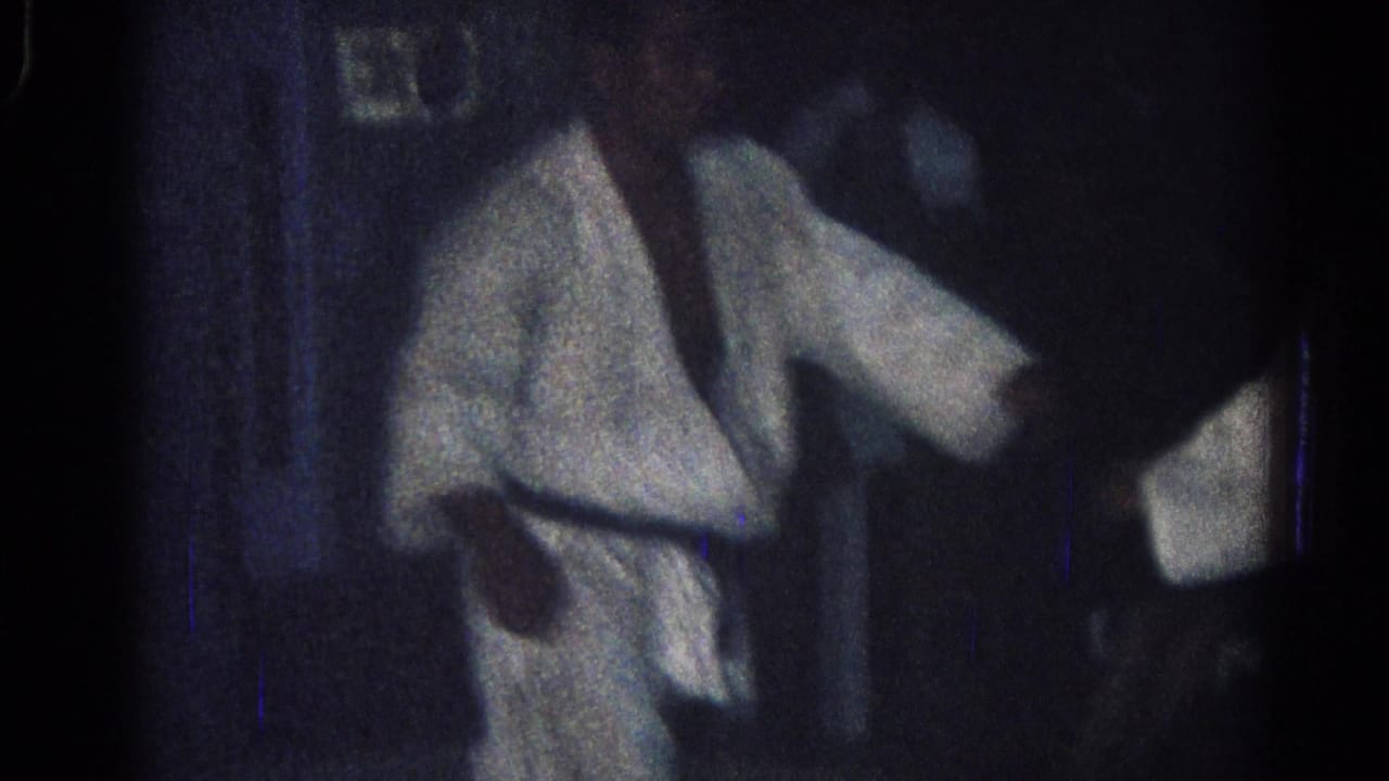 A white-clad judo practitioner stands near another figure during the San Gabriel practice footage, with faces softened by low-light 8mm grain.