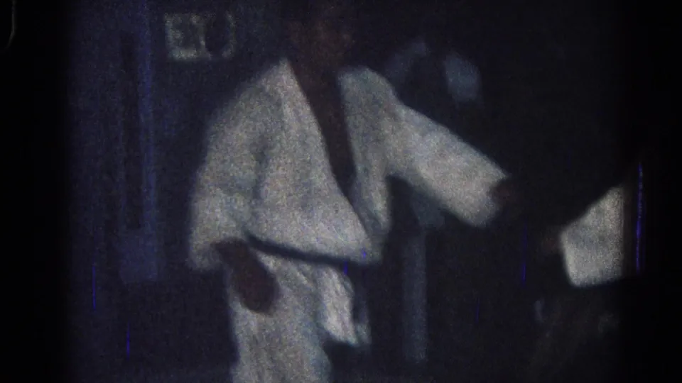A white-clad judo practitioner stands near another figure during the San Gabriel practice footage, with faces softened by low-light 8mm grain.