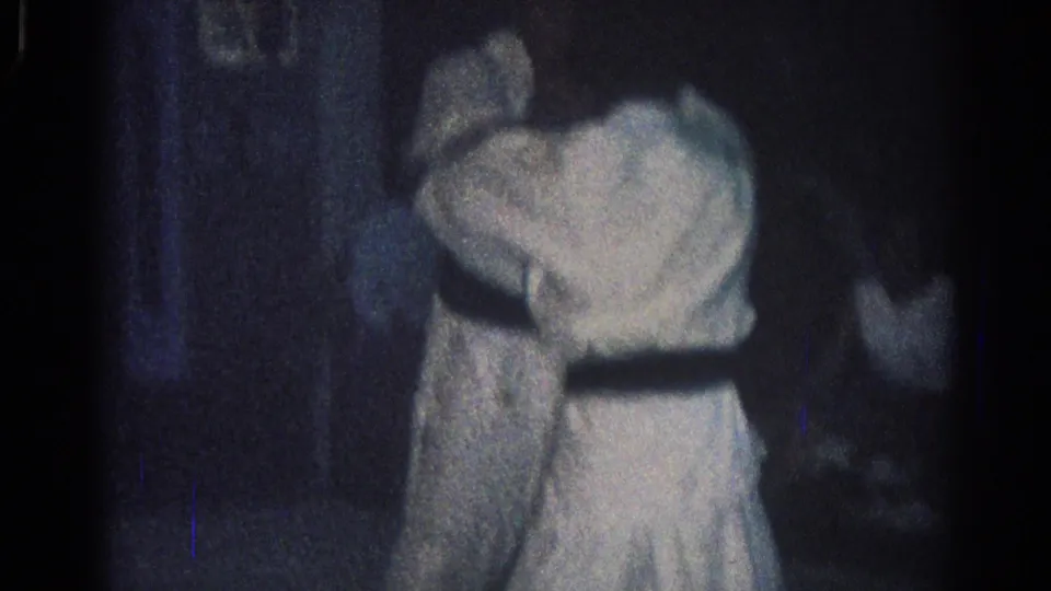 Two judo practitioners in white gi stand close in the dim San Gabriel practice room, with a partly visible person near the edge of the frame.