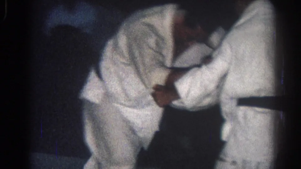 Two men in white gi bend into a grip or throw attempt during the 1966 San Gabriel judo practice, seen through grainy 8mm low-light footage.