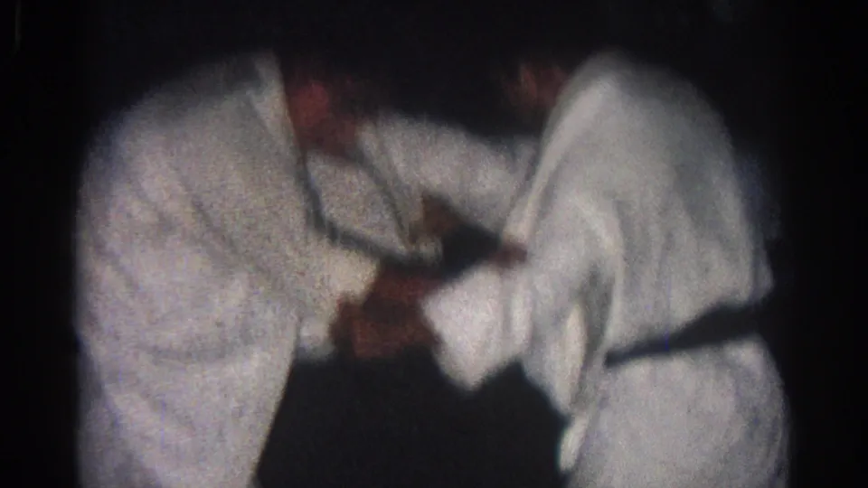 In 1966 San Gabriel home-movie footage, two adult men in white judo gis begin a close grappling exchange on a dark mat.