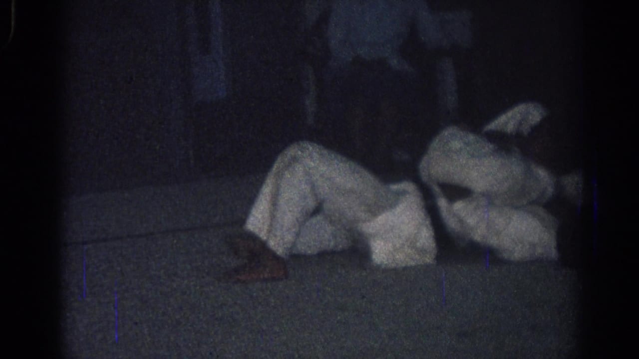 A person in a white uniform lies on the floor with another figure partly visible nearby.