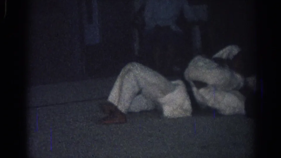 Dim 1966 San Gabriel training-room frame with a martial arts practitioner on the floor during a grappling exercise.