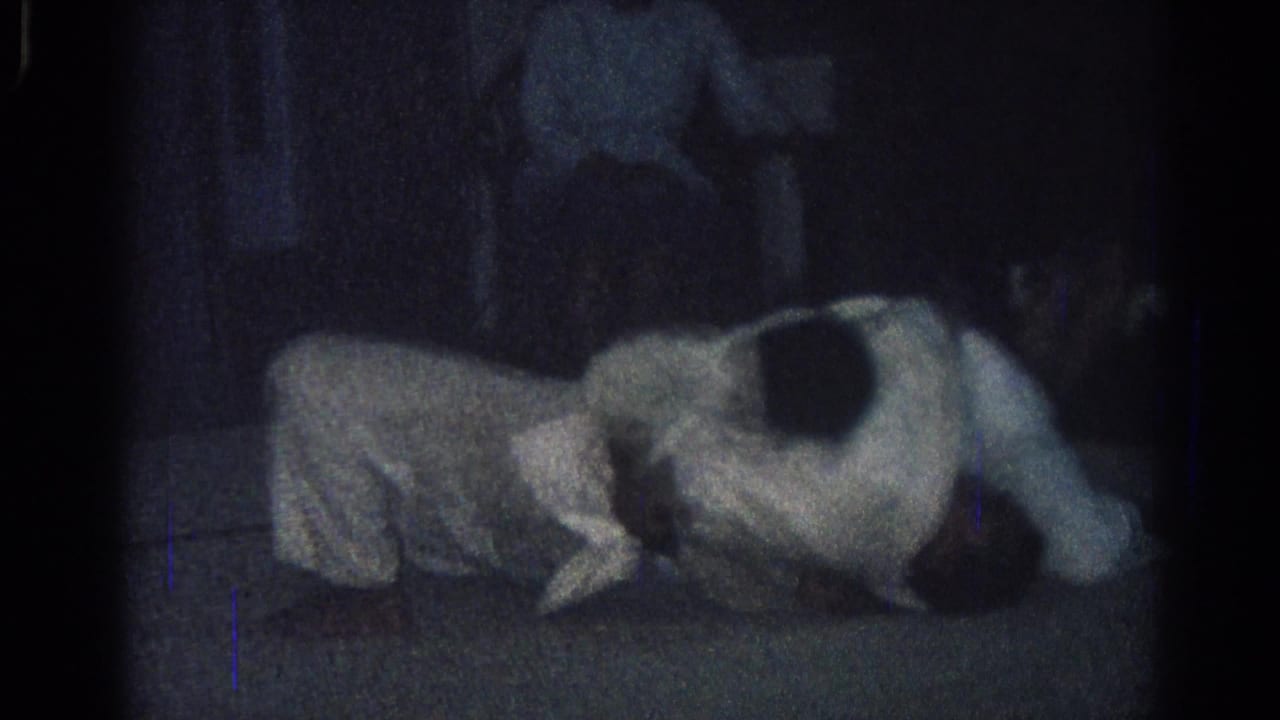 Two men in white uniforms grapple on a dark mat with observers nearby.