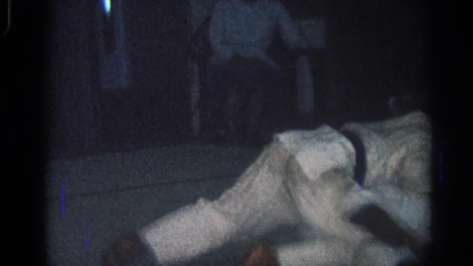 San Gabriel, California 1966 frame of two men in white uniforms continuing a ground grapple on a dim practice mat with spectators behind them.