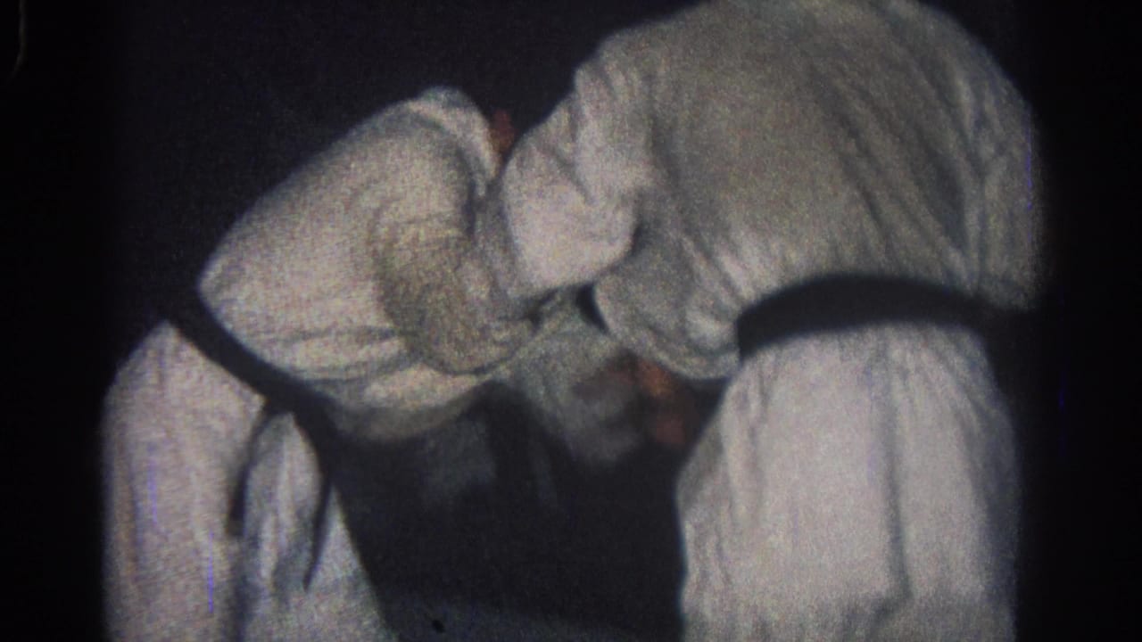 Two martial artists in white gi and black belts bend forward while grappling in a dark indoor space.