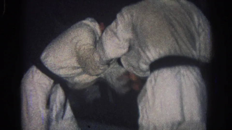 1966 San Gabriel home-movie frame of two gi-clad martial artists leaning into a close grappling exchange under dim indoor light.