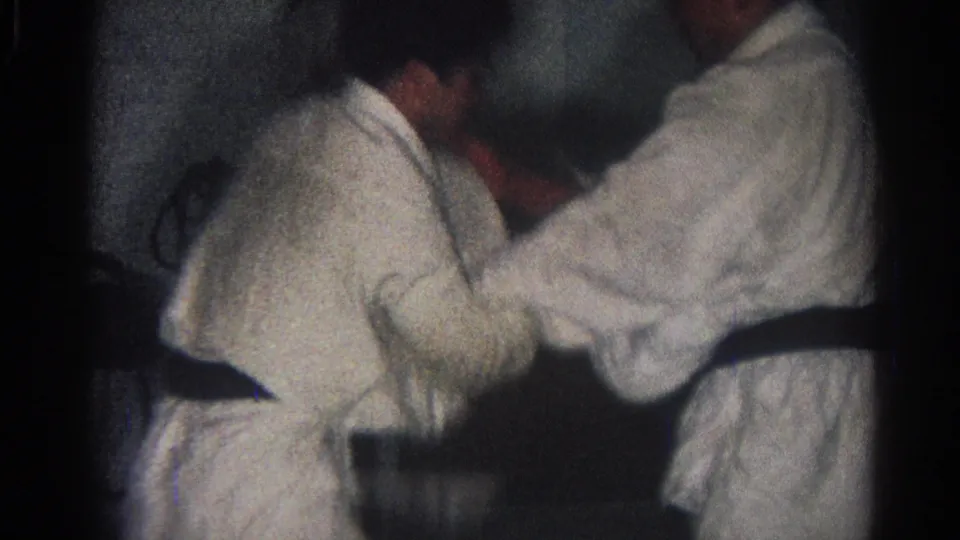 Close handheld 1966 San Gabriel frame of two black-belt martial artists locked in a standing grip during an indoor match or demonstration.