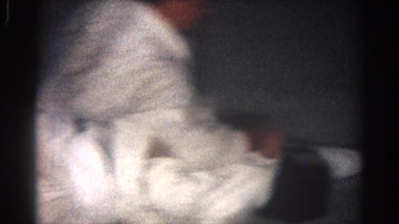 A soft, low-light 8mm frame shows a white-uniformed martial artist bending during close-contact ground practice.
