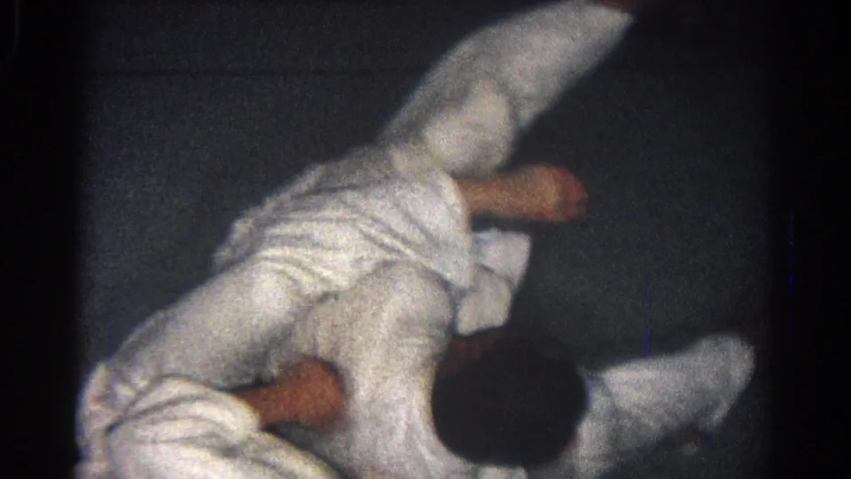 Two adult male martial artists in white gi uniforms grapple on a dark mat in 1966 San Gabriel home-movie footage.