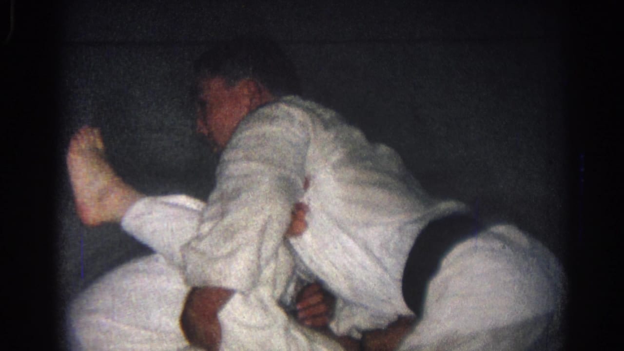 A male martial artist in a white gi appears to control another fighter on the mat during 1966 training footage.