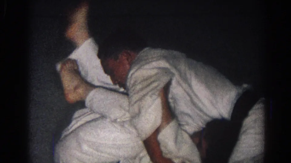Two adult men in white martial arts uniforms continue a close floor grappling exchange in San Gabriel archival footage.