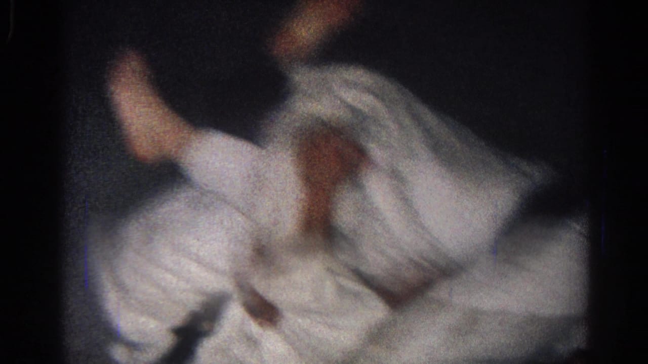 Two men in white gi uniforms grapple low to the ground in a dark room.