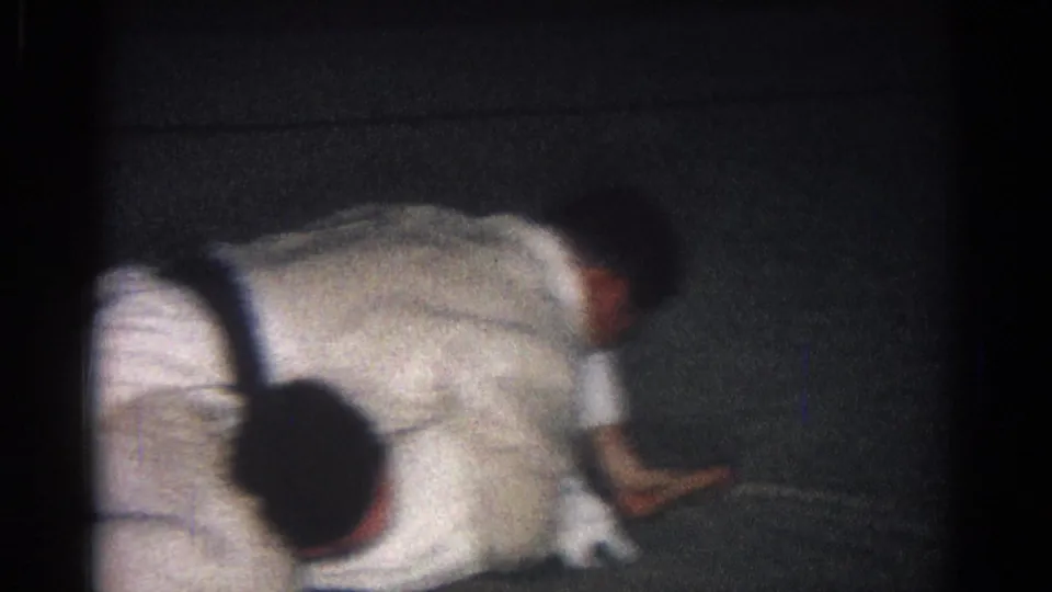 Two adults in white martial arts uniforms continue ground grappling on a dark mat in the 1966 San Gabriel clip.