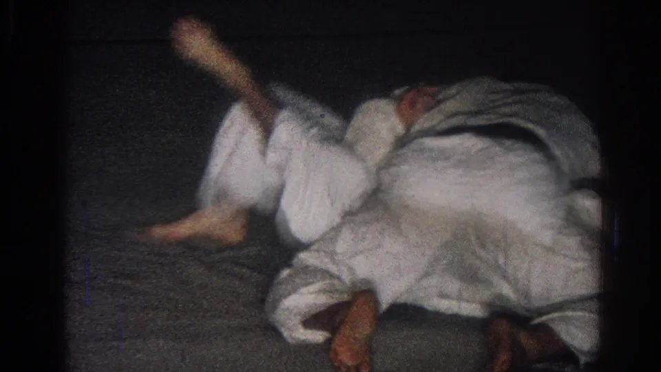 1966 San Gabriel 8mm frame showing two men in white martial arts uniforms grappling low on a dark indoor mat.