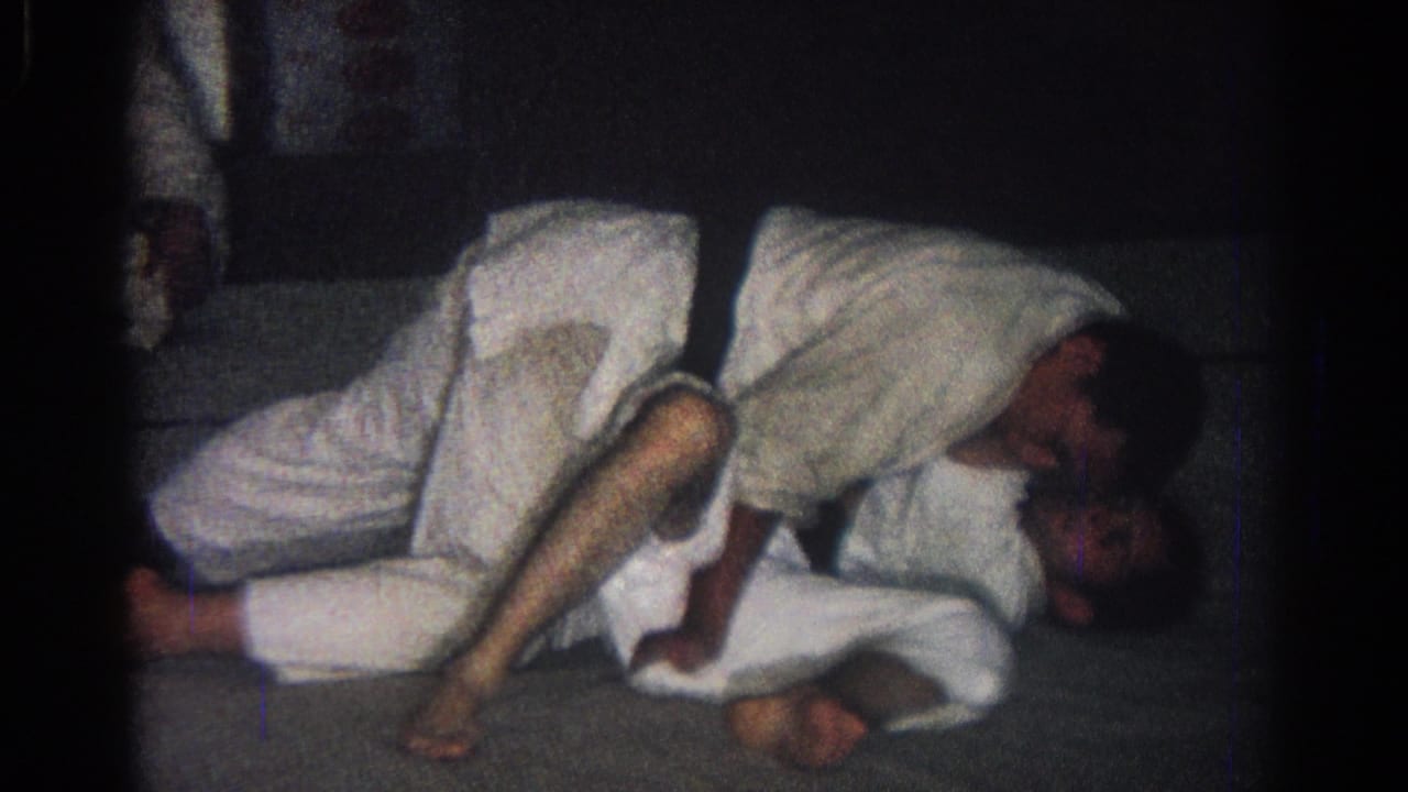 Two people in white gi grapple on a mat while another person stands nearby.