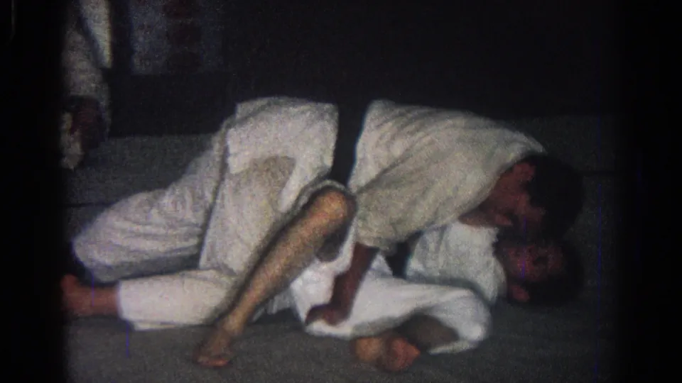 1966 San Gabriel home-movie frame showing two judo students in white gi practicing a ground technique on a mat as another student observes.