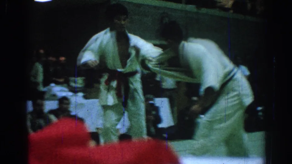 Two competitors in white gi grapple on a mat during a 1966 San Gabriel judo match, with spectators visible behind them.