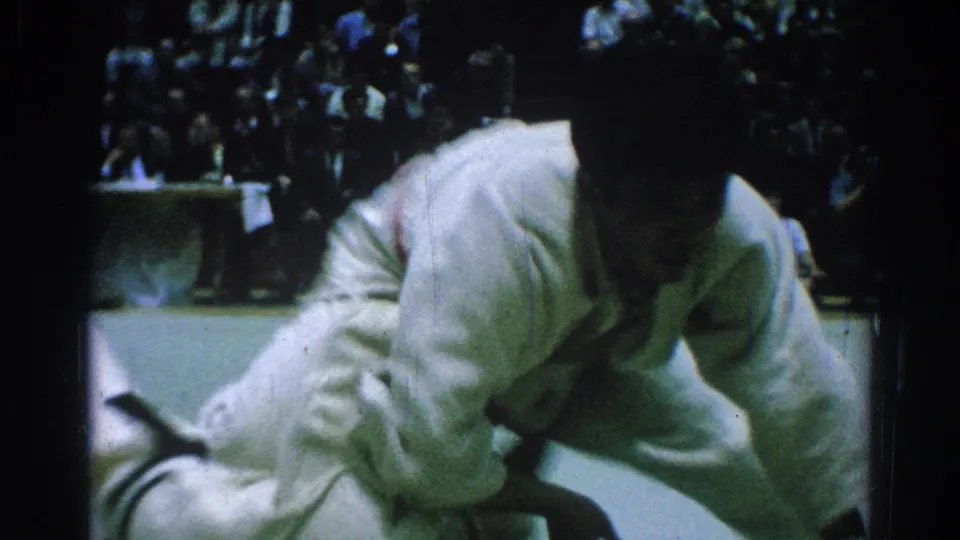 A competitor in a white gi drops low during the judo exchange, with spectators and another athlete partly visible in the dark background.